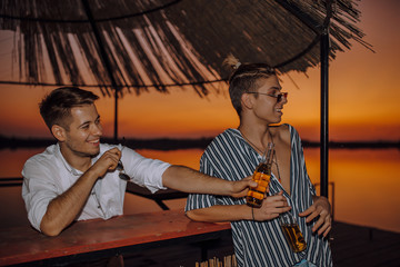 Obraz na płótnie Canvas Two young friends drinking beer and pointing with hand to someone while enjoying in a good mood on the beach bar