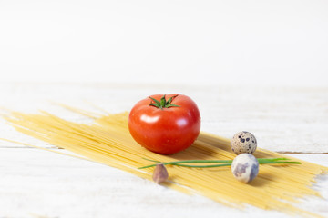 Tomato laying on bunch of spaghetti with quail eggs, scallions, garlic on white wooden background