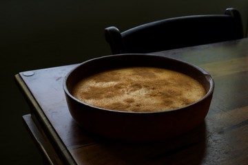 Dark food photograph of a milk tart topped with cinnamon in an earthen bowl on the corner of a wooden table.  The top of a wooden chair is seen in the background.