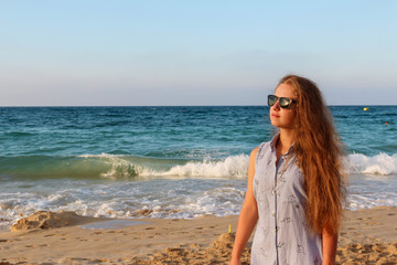 a beautiful young tourist girl with long brown unpainted hair and glasses stands on the beach against the background of the blue sea and Golden clean sand