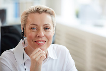Portrait of smiling blonde woman wearing headset and looking at camera while working in support...