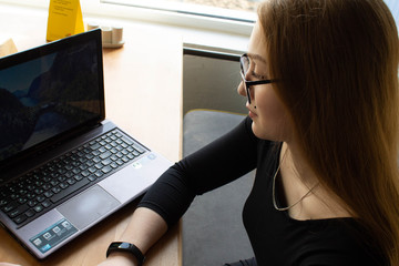 Beautiful sexy girl in a black dress and glasses sits at the table and works at the computer. Distance learning. Remote work.