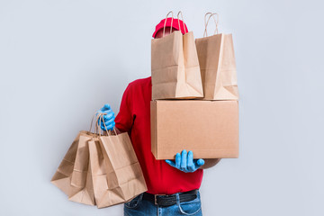 The concept of safe delivery. A courier in a red uniform and protective mask and gloves holds a large order, a cardboard box and many paper bags, contactless delivery of orders in quarantine.