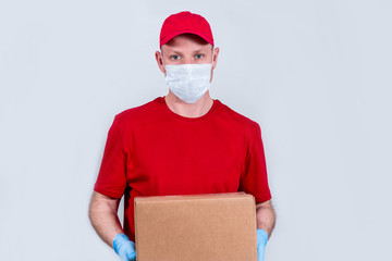 Fototapeta na wymiar The concept of safe delivery. A courier in a red uniform and protective medical mask and gloves holds a cardboard box, contactless delivery of orders in quarantine during the coronavirus pandemic.