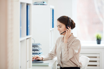 Side view portrait of smiling businesswoman speaking to microphone while working with laptop in...