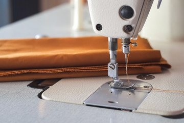 Needle presser foot with thread of white industrial sewing machine with brown cloth close-up.