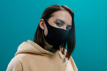 Fashionable girl in black medical mask and beige hoodie looks into the frame