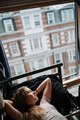 Young woman with long light brown hair wearing a nude short-sleeved shirt is lying on a bed overlooking the city.