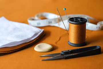 Needle and pin on thread rolls for hand sewing on brown cloth with scissors cut fabric and sketch yellow chalk with tape measure close-up.