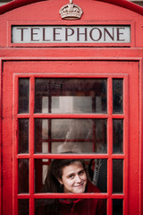 Portrait of a young woman inside a typical London red phone booth with her face resting on the glass on a clear autumn day.