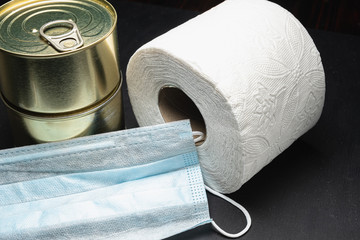 roll toilet paper and two cans of canned food, and used medical mask coronavirus virus protection. horizontal orientetion. dark background.