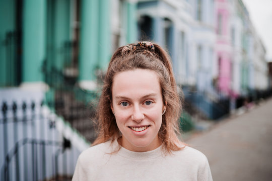 Portrait of an attractive natural young girl smiling with blue eyes, blonde hair and nude sweater, on a street with colorful houses in London.