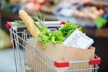 Background image of shopping cart with fresh groceries, focus on shopping list in paper bag, copy space
