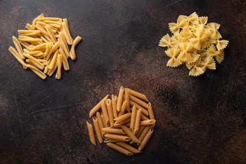 various types of uncooked pasta (treccia, farfalle, whole grain penne) on a dark concrete background. Italian Cuisine. top view.