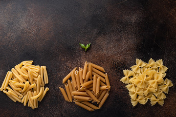 assorted uncooked pasta (treccia, farfalle, whole grain penne) on a dark background. food frame. flat lay.