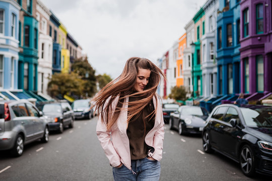 Happy young woman with long light brown hair and a pale pink jacket, is laughing through the middle of the street in London.