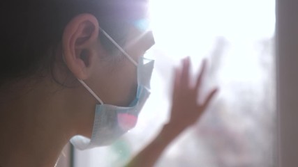 little girl looks out the window sad in a medical gauze mask. concept pandemic virus covid 19 self-isolation infection doomsday. the girl touches the glass sunlight wants to go outdoors lifestyle - Powered by Adobe