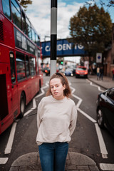 Portrait of a young woman dressed in pastel colored fall clothing and leaning against a road sign. In part of the photograph you can see a typical red double-decker London bus.