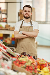 Fototapeta na wymiar Vertical waist up portrait of bearded man standing with arms crossed and looking at camera while selling fresh fruits and vegetables at farmers market
