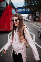 An attractive young woman with long brown hair, sunglasses and a pink coat smiles as she walks between two lanes of vehicles. In the background you can see a typical London bus.