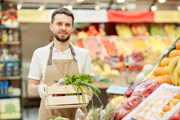 Waist up portrait of bearded farmer holding box of vegetables while selling fresh produce at market stand, copy space