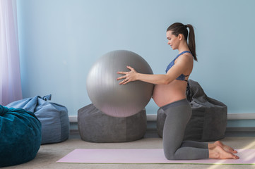 Fototapeta premium Pregnant european woman performs exercises with fitness ball. Expectant mother is doing yoga in the 3rd trimester. Girl in the classroom in preparation for childbirth.