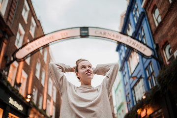 Young woman with her arms behind her head placed under a sign in a famous and colorful neighborhood in London on a cloudy autumn day.