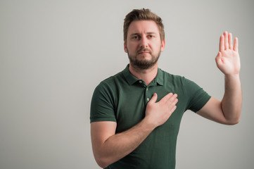 Portrait of serious stylish attractive man with thick beard, dressed in casual green t shirt taking oath gesture