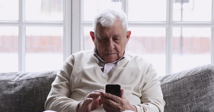 Smiling elder 80s adult grandfather learn mobile phone app sit on sofa, happy 70s senior man hold smartphone texting messages in social media reading news, old person using tech gadget concept at home