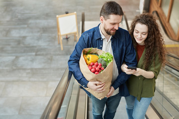 High angle portrait of happy modern couple holding bag with groceries while going up escalator in mall, copy space