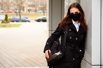 Portrait of a pretty caucasian brunette girl on a city street in a medical black mask while walking outdoors in a black coat on the background of the stairs