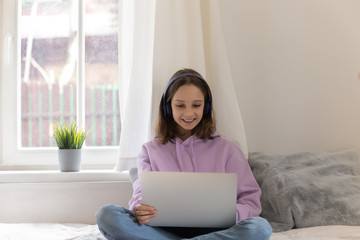 Happy teen girl wearing headphones watching videos on laptop sitting on bed in her room. Smiling...