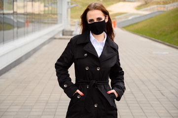 Obraz premium Portrait of a caucasian brunette girl on a city street in a medical black mask while walking outdoors in a black coat on the background of the road near the building