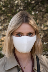 Young blonde girl in a protective mask for spreading of disease virus SARS-CoV-2. Epidemic. Coronavirus. Covid-19. Trees on the background.