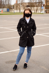 Portrait of a caucasian brunette girl on a city street in a medical black mask while walking in open air in a black coat in the background of a parking lot