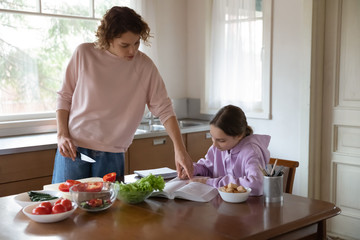 Mom helping teenage daughter with studies while cooking at kitchen table. Young adult parent mother and teen child girl doing everyday activities enjoying family casual lifestyle at home together.