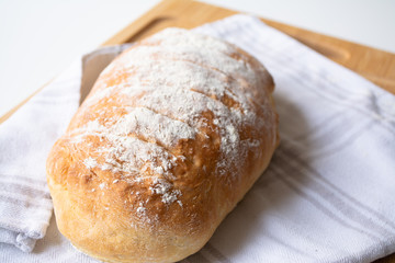 A loaf of homemade fresh bread with flour sprinkled on top in a kitchen towel on wooden cutting board with slices