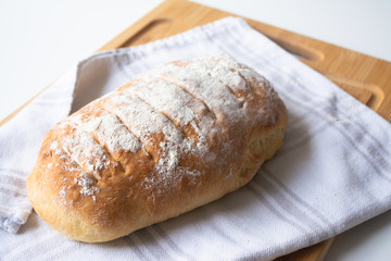 A loaf of homemade fresh bread with flour sprinkled on top in a kitchen towel on wooden cutting board with slices