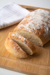 A loaf of homemade fresh bread with flour sprinkled on top in a kitchen towel on wooden cutting board with slices