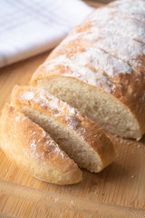 A loaf of homemade fresh bread with flour sprinkled on top in a kitchen towel on wooden cutting board with slices