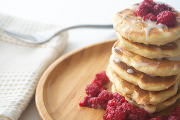 Fluffy pancakes with condensed milk and raspberries on a wooden plate with a kitchen napkin and a fork 