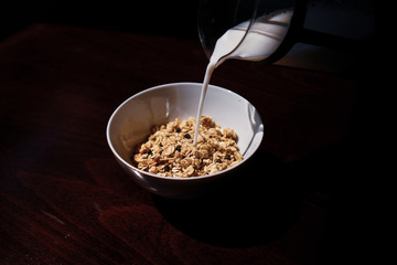 Pouring the milk into oatmeal in a white porcelain bowl on the wooden background.