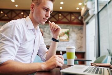 Concentrated handsome young businessman sitting at cafe table and reading important document on laptop screen
