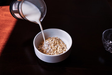 Pouring the milk into oatmeal in a white porcelain bowl on the wooden background.