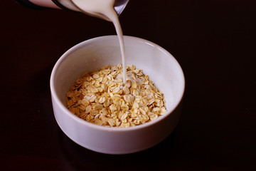 Pouring the milk into oatmeal in a white porcelain bowl on the wooden background.