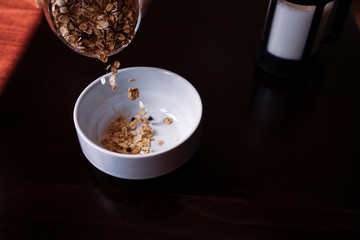 Pouring the oatmeal into the white porcelain bowl standing on wooded table.
pouring oatmeal for breakfast in a white porcelain oatmeal bowl. preparing for the breakfast with oatmeal.