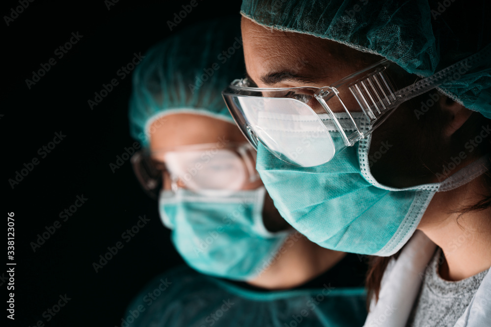 Wall mural Close of two side female and male doctors working wearing medical surgical mask, glasses, medical cap and virus protective clothing on black background with copy space. 