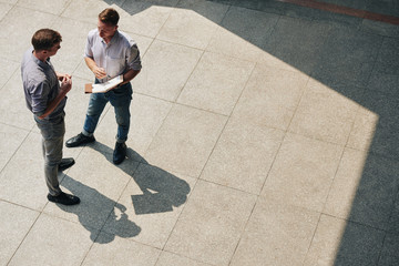 Young businessman with opened planner standing outdoors and talking to colleague about work, view...