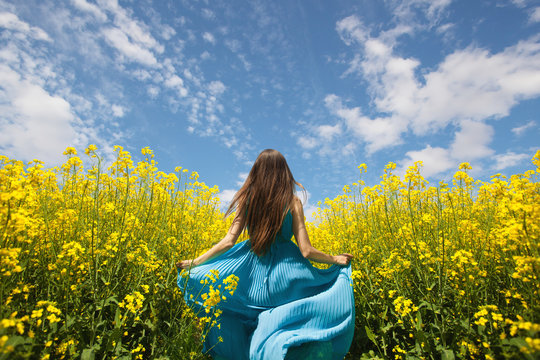 A young girl runs in a puffy blue dress on a background of a blooming field and a beautiful blue sky.