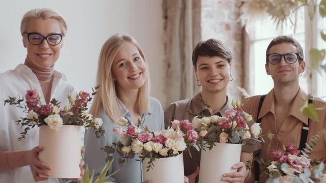 Waist up shot of happy mixed aged women and young man in apron holding beautiful flower compositions in hatboxes, looking at camera and smiling while posing on floristics masterclass in workshop